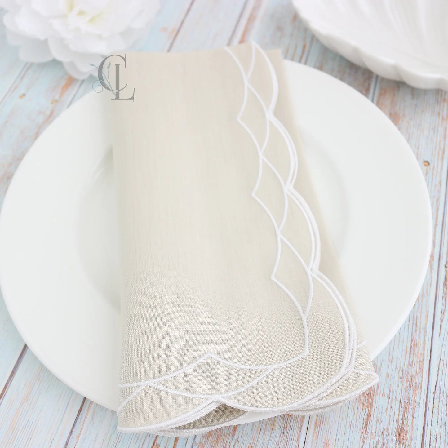 White plate with gold cutlery on a white background, featuring a Fog Beige folded scalloped dinner napkin and the brand 'Cora's Linens'.