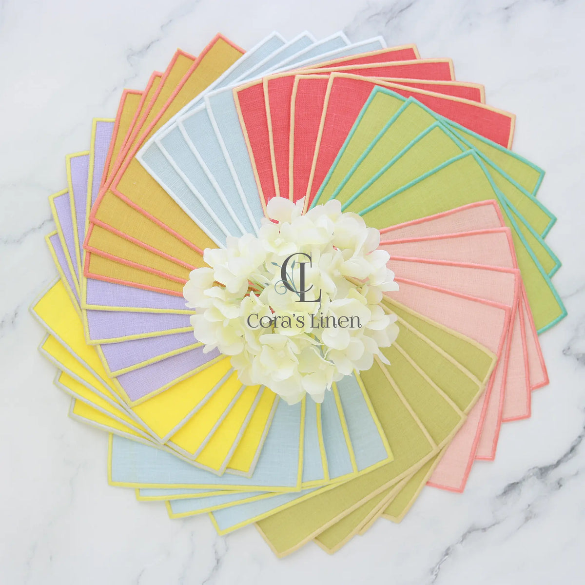 Turin cocktail napkins arranged in a circular pattern with 'Cora's Linen' branding on a marble background.
