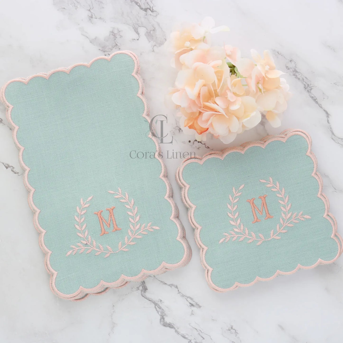 Two sets dusty blue cocktail napkins with peach monogram on a marble surface with flowers, made by Cora's Linen