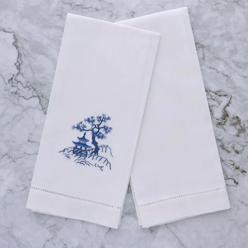 embroidered design of a tree and pagoda on a white linen guest towel next to a plain white guest towel