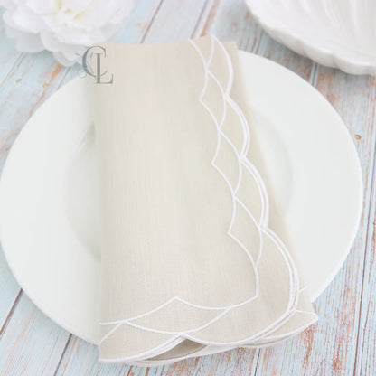White plate with gold cutlery on a white background, featuring a Fog Beige folded  scalloped dinner napkin and the brand 'Cora's Linens'.