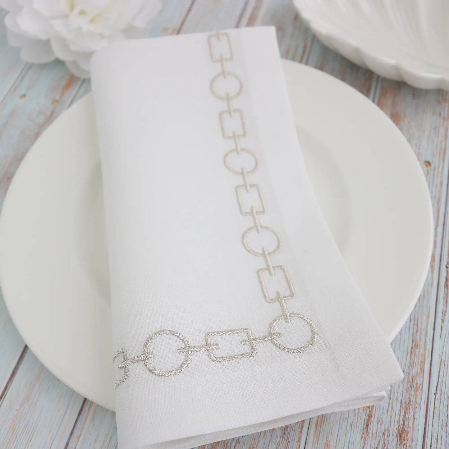 mixed chain motif embroidered in silver metallic on a white napkin, folded on a white plate, handmade by Cora's Linen