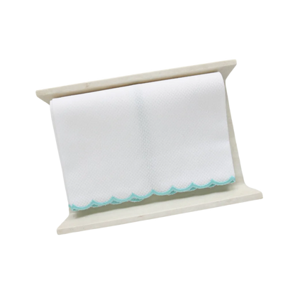 A white pique fingertip towel with an aqua blue scalloped edge, folded to fit on a cream stone towel holder