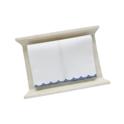 A white pique fingertip towel with a dark blue scalloped edge, folded to fit on a cream stone towel holder