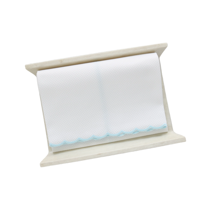 A white pique fingertip towel with a pale aqua blue scalloped edge, folded to fit on a cream stone towel holder